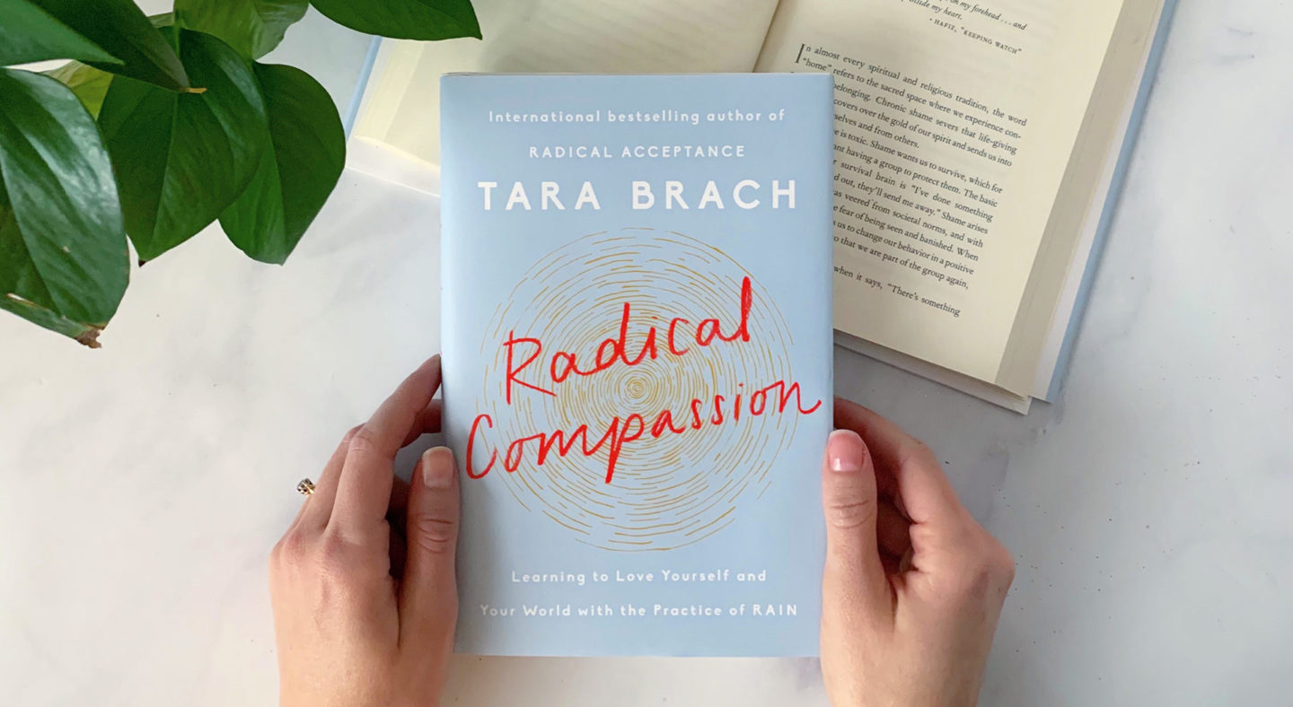 Two hands holding Tara Brach's book "Radical Compassion." Cover is light blue with title in red. Cover is light blue
